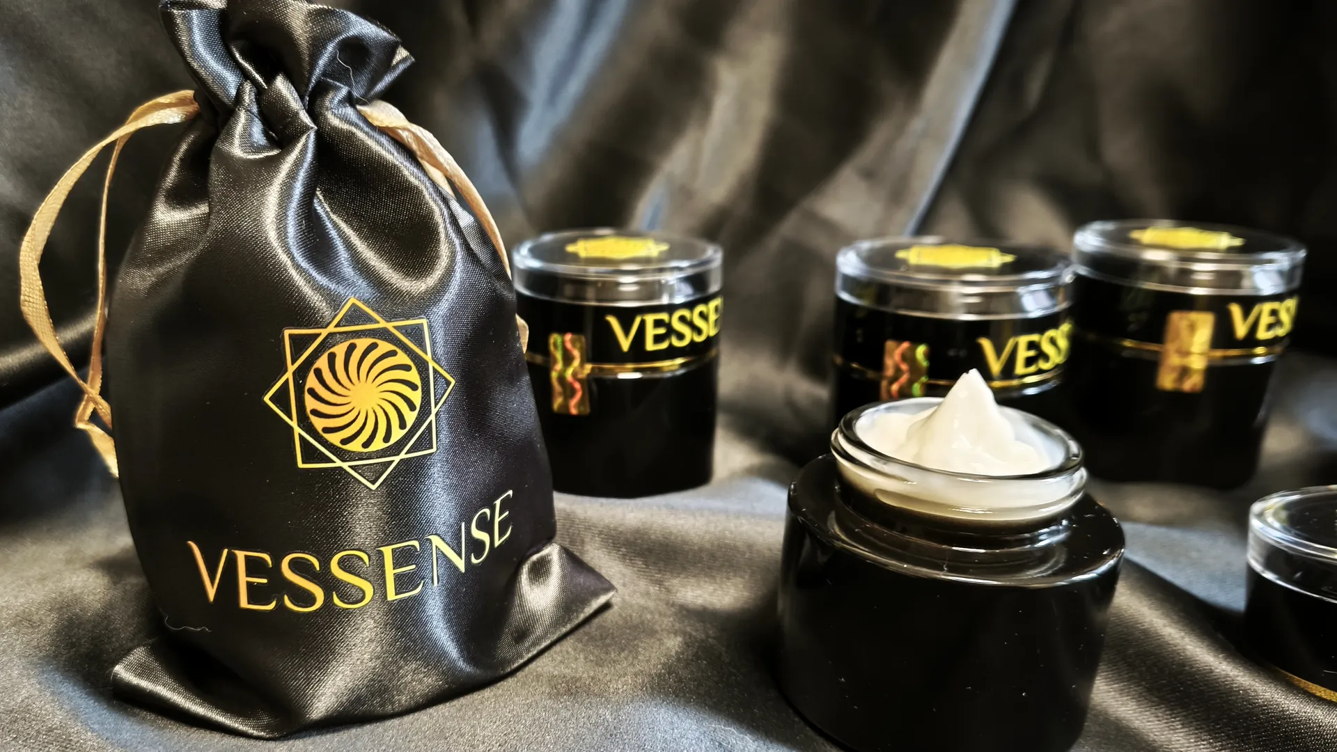 VESSENSE-Kollektion auf schwarzem Satin — Satinbeutel, versiegelte Tiegel und ein geöffneter Cremetiegel